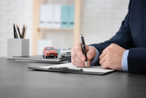 A businessman in a suit writes on a clipboard, with miniature cars and stationery on a sleek desk in a modern office setting.