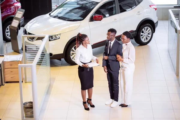 A car dealership interior featuring a sales associate discussing options with two customers, while a white SUV is prominently displayed in the background.